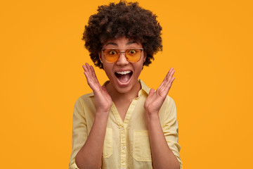 Optimistic dark skinned woman has overjoyed expression, keeps mouth opened, exclaims with surprisement, gestures with hands, poses against yellow background. African female recieves good news