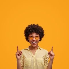 Portrait of glad African American female with joyful expression, keeps fore fingers raised, advertises something, smiles happily, isolated over yellow backgroud. Copy space for your promotional text