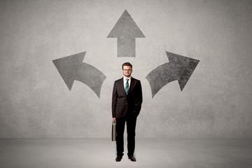 Charming businessman in doubt, choosing from three directions in front of a grey wall
