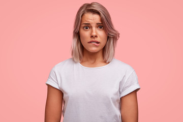 Portrait of discontent female curves lips, feels nervous before making inportant decision, has frustrated expression, wears casual white t shirt. People, emotions, facial exressions concept. - obrazy, fototapety, plakaty