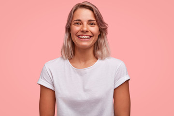 Joyful teenage girl with dental braces on teeth, rejoices positive news, wears casual t shirt,...