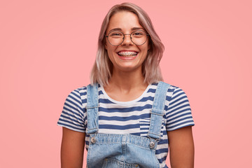 Positive glad smiling female teenager with braces on teeth, wears stylish denim overalls and...