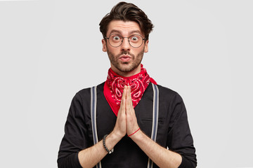 Pleadig hipster male in stylish clothes, keeps palms pressed together, has fashionable haircut and...