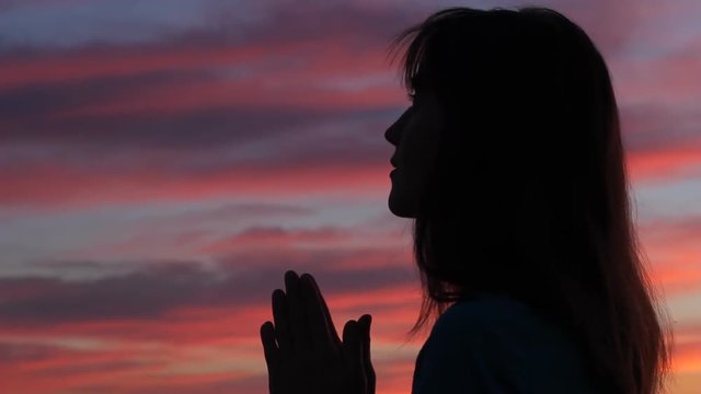 Woman with belief looking up. Silhouette profile of young woman praying with hands together to sunset sky
