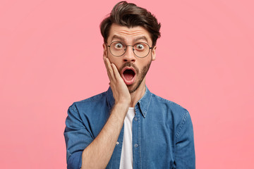 Surrprised hipster guy hears fake news or unexpected rumor, stares at camera with jaw dropped out, wears fashionable jean jacket, isolated on pink background. Amazed young man has some problems