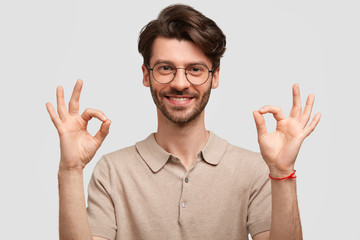Cheerful delighted male student makes ok sign, being satisfied with results of passed exam, wears...