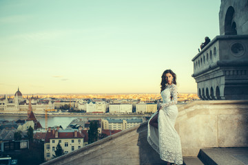 Girl with glamour look. Woman in white wedding dress on city view, fashion. Sensual woman with long hair on balcony, beauty. Bride with makeup. Fashion model style and hairstyle