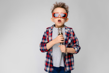 A handsome boy in a plaid shirt, gray shirt and jeans stands on a gray background. A boy wearing sunglasses. Red-haired boy sings into the microphone