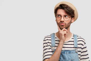 Pensive bearded male gardener looks thoughtfully aside, thinks about reap harvest, wears striped casual sweater, denim overalls and straw hat, poses against white background with copy space.