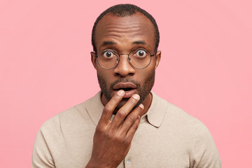 Close up shot of handsome stupefied young African American male stares surprisingly as reads strange headline in journal, isolated over pink background. Black hipster male realizes hoax or fraud