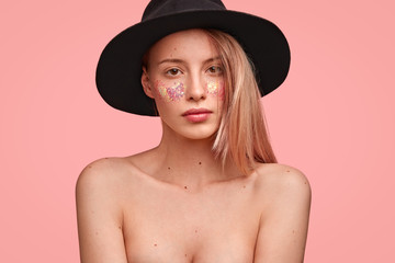 Lovely naked female with soft healthy skin, wears black hat, poses against pink background, has glitter on cheeks, pleasant appearance. Beautiful nude young woman poses in studio, shows her beauty