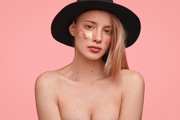 Headshot of beautiful female with naked body, demonstrates her natural beauty, has glitters on cheeks, wears only fashionable hat on head, poses over pink background. Sparkling skin. Fashion concept