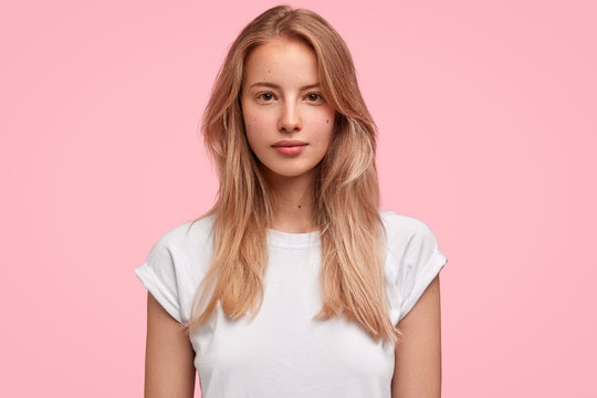 Horizontal portrait of serious beautiful female with long hair, has no make up, looks delightfully at camera, wears casual white t shirt, poses against pink background. People, lifestyle, beauty