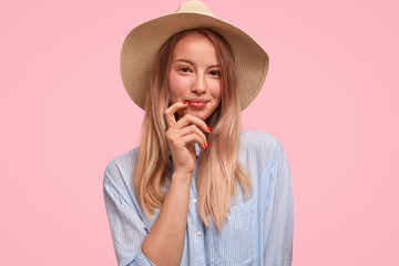 Isolated shot of elegant gorgeous lady in big summer hat and casual striped shirt, enjoys vacation and holidays, travels abroad, looks delightfully at camera, isolated over pink studio background.