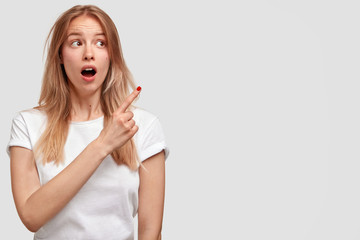 Lovely surprised female with appealing look, has long hair and shocked expression, indicates aside at blank copy space, wears casual white t shirt. Human facial expressions and advertisement concept