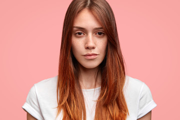 Obraz premium Headshot of pleasant looking young European woman with freckled healthy skin, wears casual white t shirt, being deep in thoughts, demonstrates her natural beauty, isolated on pink background.