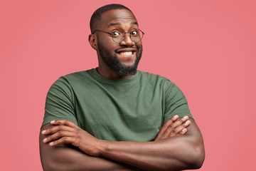 Photo of attractive dark skinned male with glad expression, keeps hands crossed, rejoices achieving success in particular sphere, wears casual t shirt, round spectacles, isolated on pink background