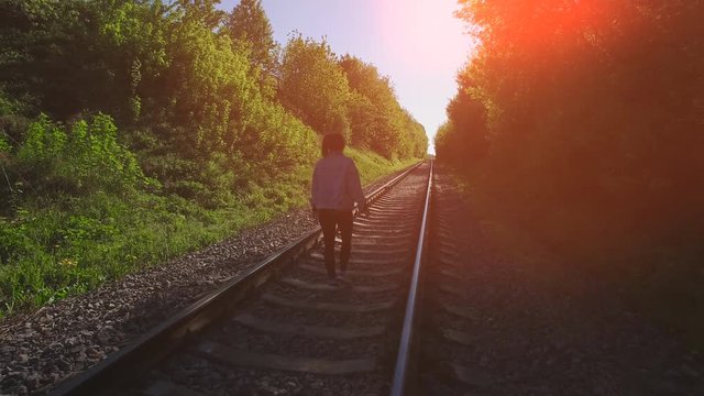 Young girl are walking on the railways. She looking forward and go ahead. Summer sunset with orange patch of reflected light. Funny and dangerous jumping on the train road.
