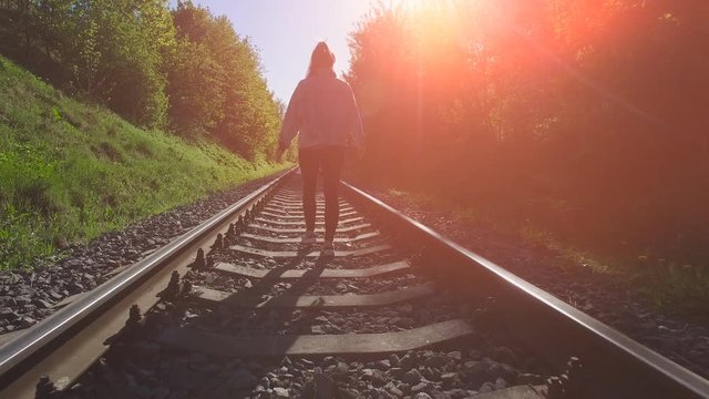Young girl are walking on the railways. She looking forward and go ahead. Summer sunset with orange patch of reflected light.