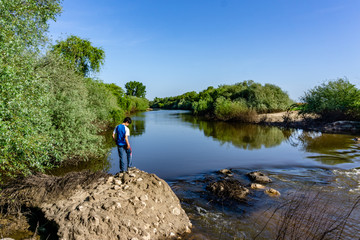 The teenager stands on top of a large stone boulder on the bank of the Sorraia River and looks at the river below. The river Sorraia in the summer - bright green vegetation,  blue sky