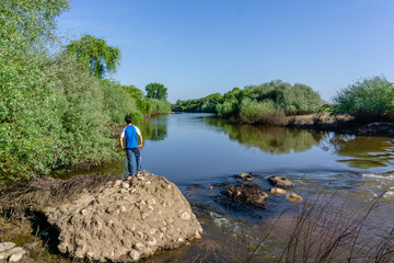 The teenager stands on top of a large stone boulder on the bank of the Sorraia River and looks at the river below. The river Sorraia in the summer - bright green vegetation,  blue sky