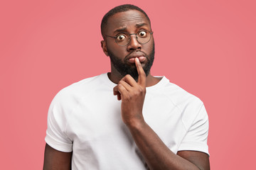 Portrait of surprised stunned African American male worker, puzzled to realize his fault, stares through spectacles, has dark bristle, healthy pure skin, wears casual t shirt, model in studio