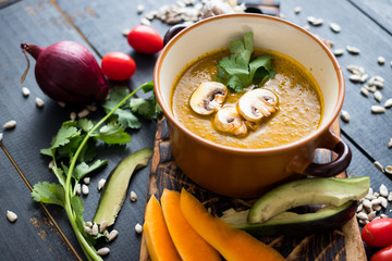 Raw vegan pumpkin dip soup with mushrooms, avocado, garlic, coriander, sunflower seeds, tomatoes. In bowl on wooden board. Organic farm products. Dark background. Vegetarian healthy food concept.