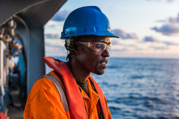 Tired Seaman AB or Bosun on deck of vessel or ship , wearing PPE personal protective equipment - helmet, coverall, lifejacket, goggles. Safety at sea