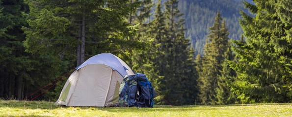Tourist camp on green meadow with fresh grass in Carpathian mountains forest. Hikers tent and backpacks at camping site. Active lifestyle, outdoor activity, vacation, sports and recreation concept. © bilanol