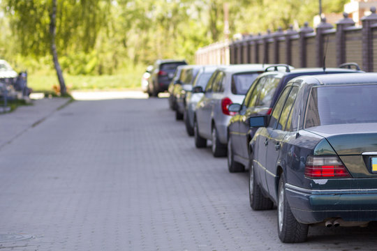 Long row of cars parked in quiet neighborhood on clean empty paved street along new stone fence on background of beautiful green trees on bright sunny summer or spring day.
