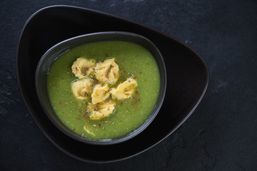 Black bowl with broccoli cream-soup and tortellini, top view on a black stone background, horizontal shot