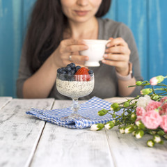 A woman is eating pudding chia with berries and drinking fragrant coffee. Romance and bouquet of pink flowers. Happy girl emotions. Woman in pajamas. Place for text.
