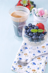 A glass of coffee and pudding chia with blueberries and strawberries for breakfast. Vegetarian healthy food. Blue background. Napkin with flowers and flowers in a vase. Bright colors. Vertical photo