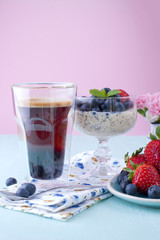 A glass of coffee and pudding chia with blueberries and strawberries for breakfast. Vegetarian food. A small bouquet of flowers, a blue and pink background.
