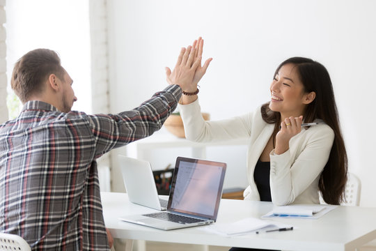 Young asian and caucasian partners giving high five at workplace, diverse motivated colleagues celebrate goal achievement, business win, successful teamwork, good result, support and victory concept