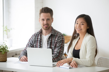 Obraz premium Smiling caucasian businessman and asian businesswoman looking at camera, diverse interns managers posing at workplace sitting in front of laptop, young motivated business professionals team portrait