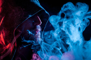A young man in a black cape smokes and exhales large clouds of colored blue and red  smoke on a...