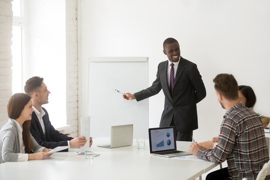 Smiling friendly african american coach in suit discussing business presentation with team people in meeting room, black ceo manager presenting new plan or project results to employees group