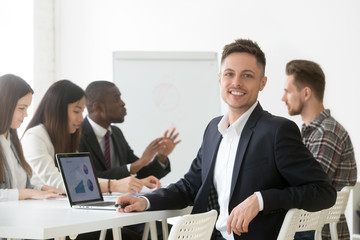 Smiling young businessman in suit looking at camera posing with laptop at group meeting, happy project manager, financial analyst, successful accountant or professional executive employee portrait