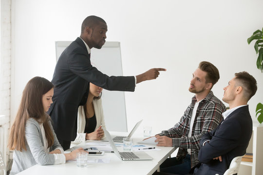 Angry rude african businessman pointing finger threatens caucasian colleague at team meeting, black executive office worker blaming partner showing disrespect, racial hate and discrimination concept