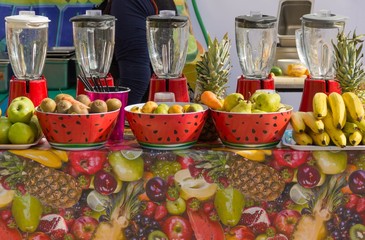 Five fruit juice makers lined up on market stand with bowls full of fruits in front