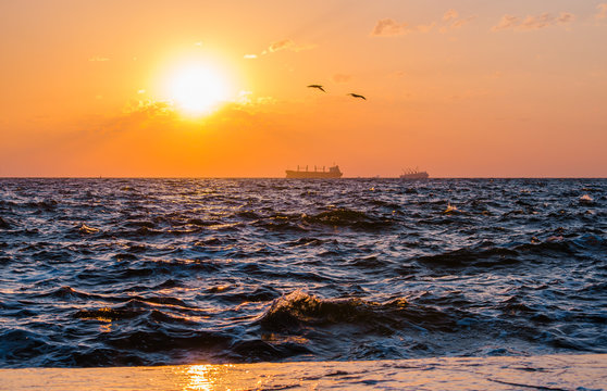 A ship sailing on the horizon at the sunrise. Sun rising over it making beautiful rays in an orange sky. Water splashes on the both sides of the photo 