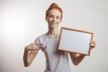 Smiling attractive red-haired female with bun pointing with finger on wooden empty frame holded in another hand isolated over white background