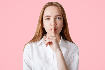 Confident lovely female with long straight hair, blue eyes and healthy soft skin, shows silence sign, asks to be silent, wears elegant white shit, isolated over pink background. Conspiracy concept