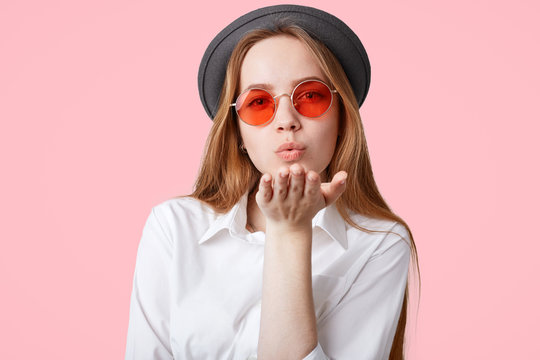 Lovely fashionable hipster girl wears trendy pink shades and black hat, blows air kiss at camera, poses against pink background, expresses her love to surrounding, Pleasant looking female flirts