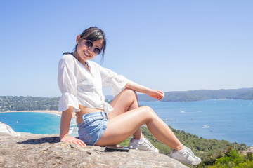 Young woman with sunglasses sits on the background of Palm beach on top of a rock at the lighthouse in Barrenjoey, Sydney, Australia.