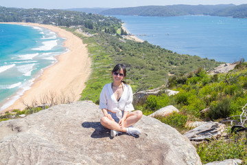 Naklejka na ściany i meble Young woman with sunglasses sits on the background of Palm beach on top of a rock at the lighthouse in Barrenjoey, Sydney, Australia.