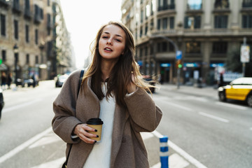 Obraz premium Portrait of charming female with take away coffee in hand standing on urban background. Beautiful young woman in a good mood dressed in stylish coat looking at camera enjoying city walk.