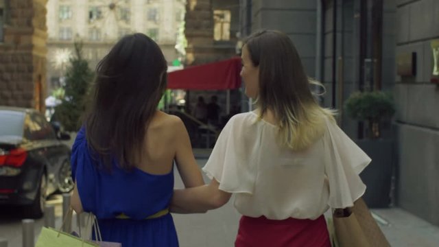Rear view of cheerful beautiful women carrying lots of colorful shopping bags and chatting while walking on the street after shooping. Joyful female friends with shopping bags walking in trade centre.