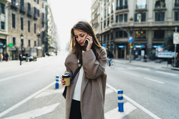Fototapeta premium Successful business woman making business calls by a mobile phone to the partners while going to office with coffee. Fashionable hipster girl wearing stylish clothes chatting with friend by smartphone